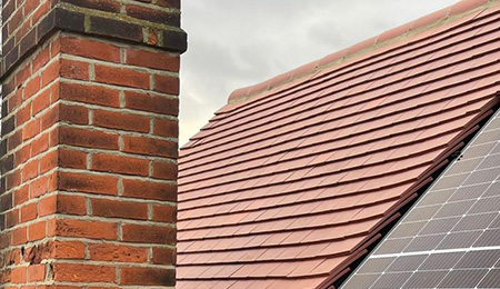 Roof with red tiles, chimney, and solar panels under a cloudy sky.